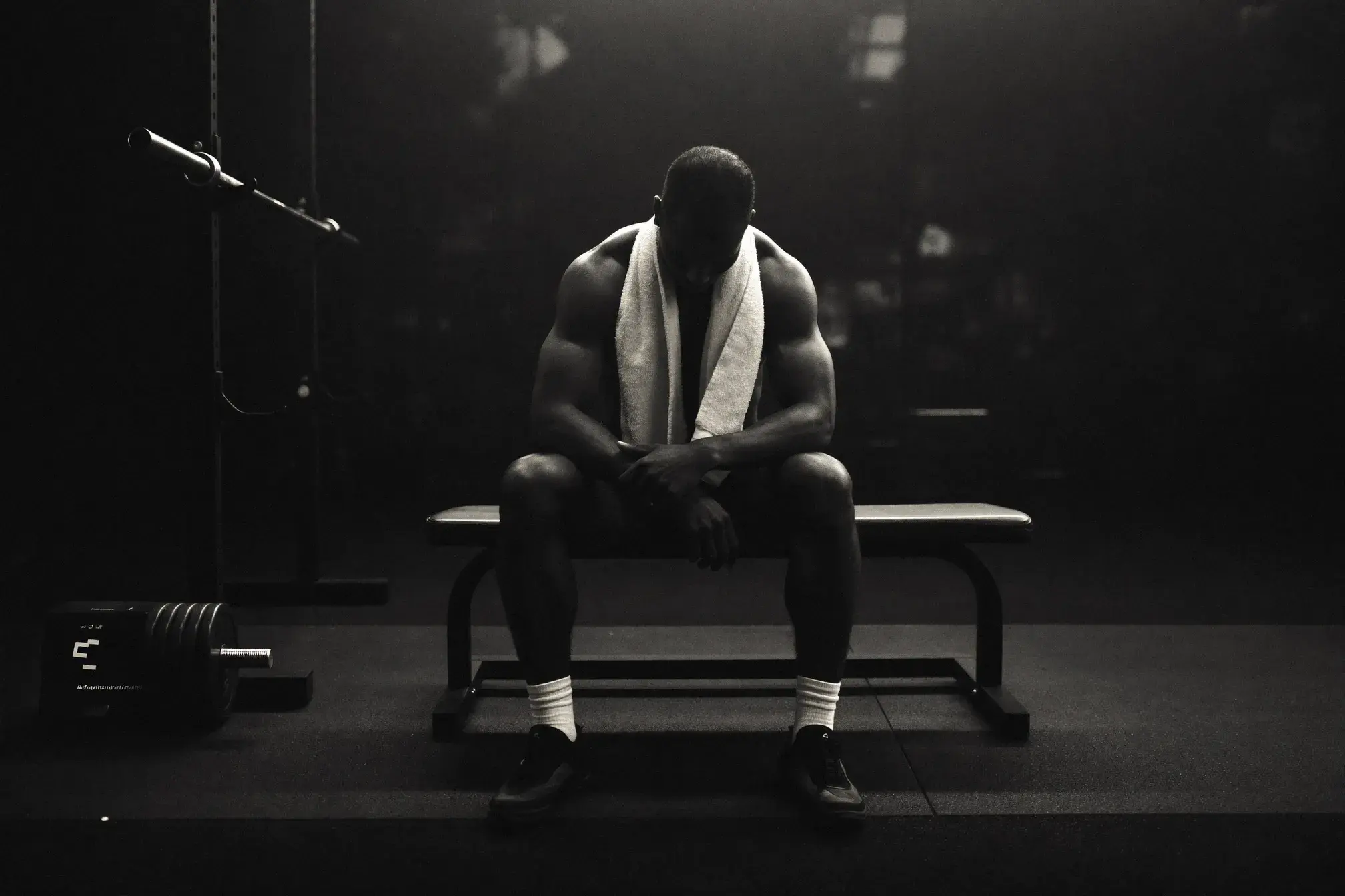 Athlete sitting quietly on a gym bench after a long training session, head bowed, towel around neck - the stillness of finishing strong