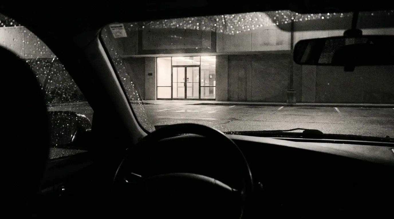 View through a rain-flecked windshield at night toward the lit entrance of an empty gym across a wet parking lot - the short distance still left to walk