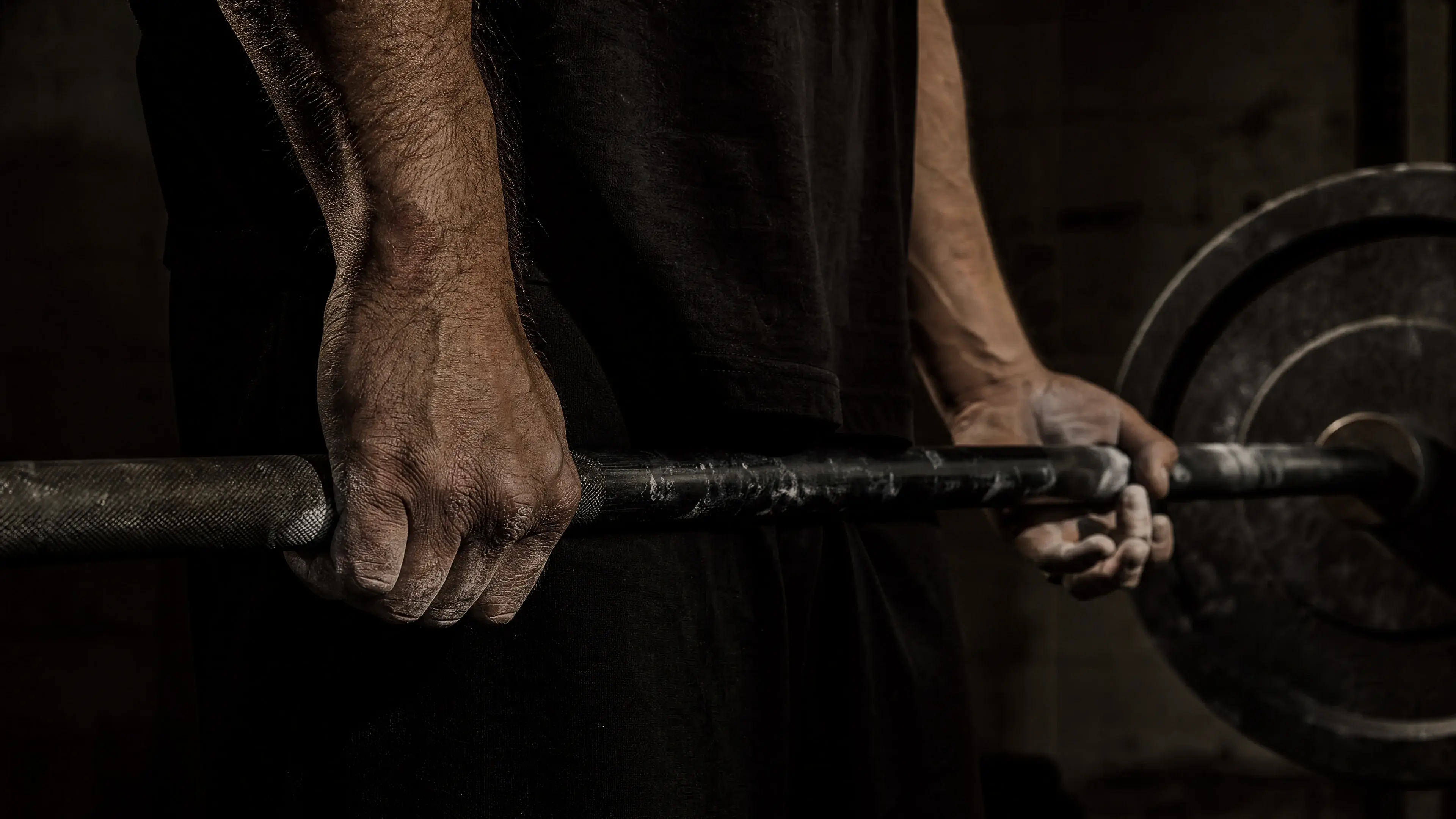 Man's calloused hands gripping a chalk-dusted barbell at lockout in a dimly lit garage gym — what the bible says about physical strength